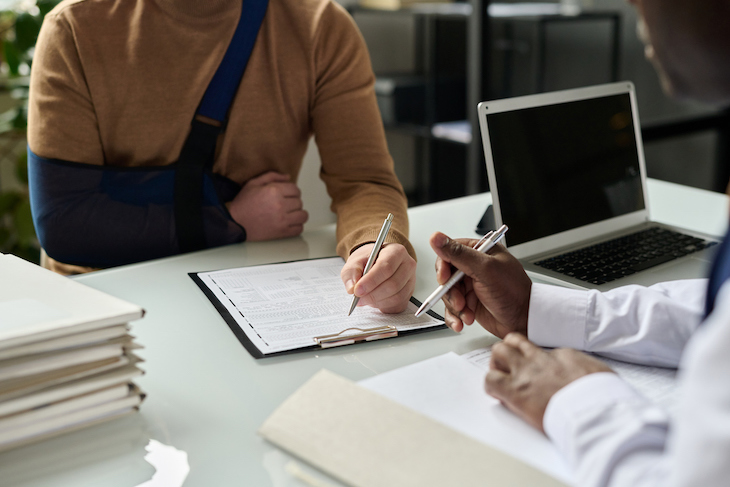 Close up of man filling in medical insurance form, injured hand in sling in background, copy space