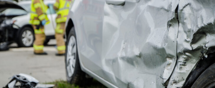 Close up on a car crashed during an accident with two firemen in background with firetruck during a day of summer